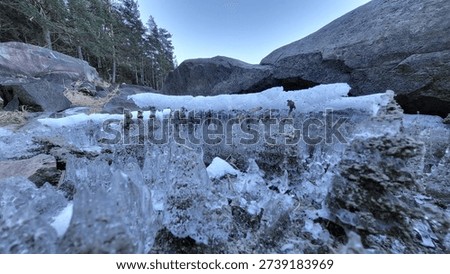 Dramatic frozen waterfall displaying massive icicles and sculptural ice formations clinging to rocky cliffs in Norway. Numerous translucent columns hang vertically from overhanging stone ledges, their cylindrical shapes varying in diameter from finger-thin to arm-thick, creating natural organ pipe arrangement. These crystalline structures formed gradually as dripping water solidified during sustained subfreezing temperatures, building layer upon layer into impressive frozen cascades. The transparency reveals internal striations documenting different freezing episodes and varying water flow rates throughout formation process. Dark weathered bedrock provides stark contrast against pale blue-white coloration of suspended frozen material. Snow caps visible atop horizontal rock surfaces while vertical faces remain exposed, showing selective accumulation patterns driven by gravity and wind. Massive boulders scattered across foreground demonstrate geological character of this rugged terrain. Dense coniferous forest visible in background distance, evergreen trees establishing geographical context within typical Nordic mountain environment. Clear blue sky overhead suggests cold stable weather conditions necessary for maintaining these delicate structures. Low-angle perspective emphasizes monumental scale of formations, making viewer feel diminutive before nature's architectural achievements. Documentary photograph from southeastern Norway captured during March when late winter conditions still produce spectacular frozen displays. Natural illumination reveals subtle color variations within crystalline material while maintaining excellent detail throughout composition. Real location shot without digital manipulation preserves authentic character of this seasonal phenomenon. Perfect for projects exploring geological themes, winter beauty, natural sculptures, environmental documentation, Nordic landscapes, or artistic collections celebrating ephemeral creations. Horizontal format provides excellent compositional balance betwe