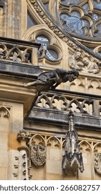 A dramatic gargoyle juts out from St. Vitus Cathedral, beneath stained glass and sculptural relief, showcasing detailed gothic stonework.