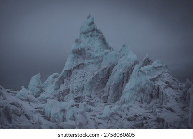 A dramatic glacial ice formation under overcast sky, showcasing rugged textures and blue-white ice peaks in a remote mountainous region.