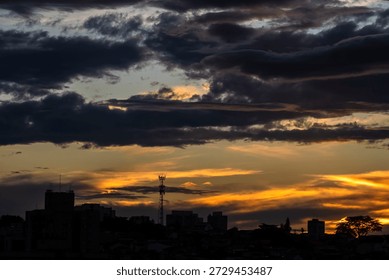 A dramatic golden sunset casts a vibrant glow over a city skyline. Dark clouds part to reveal the brilliant sun, silhouetting buildings and a lone tree against the luminous sky.