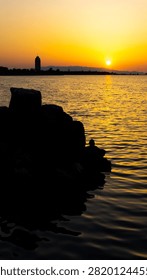 Dramatic golden sunset over a calm water surface reflecting warm sunlight, featuring dark rocky foreground silhouettes and a distant tower on a misty horizon.