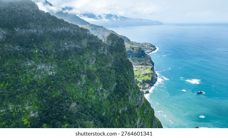 Dramatic green cliffs meet the Atlantic's turquoise waters in Madeira. Boaventura villages nestle along the rugged shoreline under a cloudy sky, offering breathtaking views.