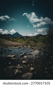 Dramatic ground-level view of muddy rocky mountain trail puddle with wild grass, sharp peaks and dramatic teal cloudy sky in background after rain.