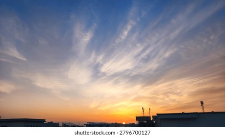 Dramatic high-altitude clouds illuminated by a colorful sunset sky. Delicate cloud formations radiate across the atmosphere above silhouetted rooftops, creating a calm and visually striking landscape. Ideal for themes of nature, weather, tranquility, evening, and atmospheric beauty.