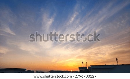 Dramatic high-altitude clouds illuminated by a colorful sunset sky. Delicate cloud formations radiate across the atmosphere above silhouetted rooftops, creating a calm and visually striking landscape. Ideal for themes of nature, weather, tranquility, evening, and atmospheric beauty.