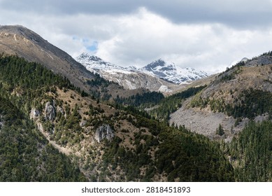 Dramatic high-altitude landscape capturing dark, forested foreground ridges giving way to jagged, snow-covered mountain peaks under a heavy overcast sky in Yunnan, China.