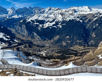 A dramatic high-altitude view looking down from the snowy ridges of Männlichen toward the alpine resort village of Wengen and the deep Lauterbrunnen Valley. The foreground prominently features massive steel avalanche protection barriers installed along the steep, snow-packed mountain slopes. These critical safety structures overlook dense evergreen pine forests and the valley floor below, highlighting alpine engineering amidst beautiful winter landscape topography.