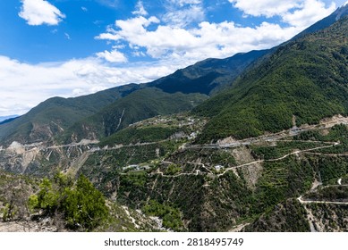A dramatic high-angle bird's-eye view looking down into a deep river canyon with zigzagging switchback roads traversing steep, emerald-green hillsides. Remote agricultural terraces and alpine settlements line the ridges in Yunnan, China.