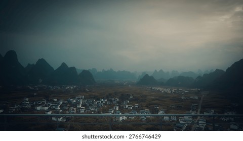 ​A dramatic, high-angle panoramic vista of the world-famous limestone karst peaks in Guilin, Guangxi Province, China. The moody, overcast sky casts a soft light over the rural village and agricultural valley nestled between the mountains. This cinematic landscape captures the iconic 