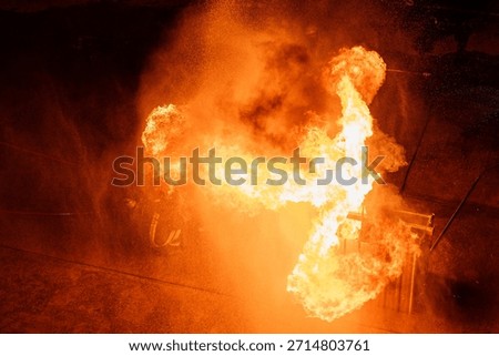 A dramatic, high-angle or top-down shot captures an enormous, turbulent mass of bright orange and yellow flames dominating the scene. The fire appears to be consuming a dark wooden-like structure or prop on the right. A firefighter wearing a yellow or light-colored helmet and gear is partially visible on the left, lying or kneeling on the ground amidst the flames and smoke, demonstrating the extreme danger and heat of the training or incident.