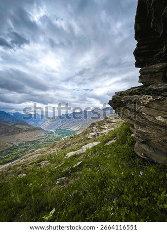 A dramatic, high-angle view of a vast, green valley with a river and a small town. The valley is surrounded by rugged mountains, with a prominent rocky cliff face in the foreground and a cloudy sky