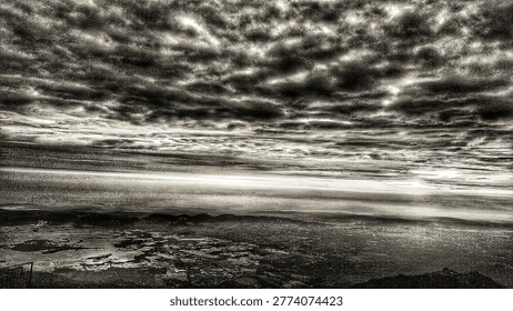 Dramatic high-contrast black and white panoramic vista from a peak, looking down over an expansive cityscape, a wide river delta, and rugged mountains under an intensely turbulent, heavy cloudscape.