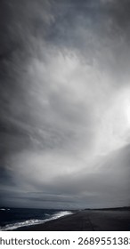 A dramatic, high-contrast monochrome image captures a vast, stormy sky dominating a dark, sandy beach with rolling waves under a hint of bright sun.
