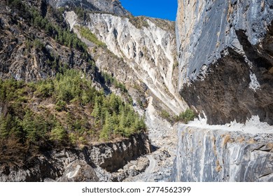 A dramatic Himalayan gorge featuring a narrow cliffside mountain road carved directly into a massive rock wall. Below, a vivid turquoise river winds through a rugged valley in Annapurna circuit Nepal