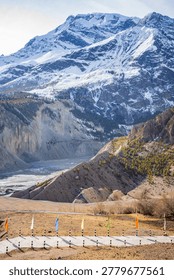 Dramatic Himalayan mountain landscape featuring towering snow-covered peaks rising above a rugged alpine valley. In the foreground, colorful Buddhist prayer flags line a mountain road