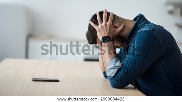 Dramatic at home, mental health problems,\
crisis, stress and expression negative emotions. Sad millennial man\
holding his head with hands, sitting at table with smartphone in\
living room interior