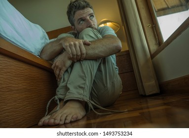 dramatic home portrait of young depressed and desperate man sitting on bedroom floor next to bed suffering depression and paranoia feeling overwhelmed and helpless
