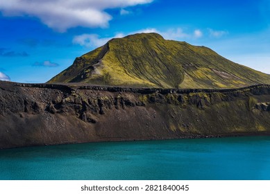 Dramatic Icelandic landscape featuring a moss-covered volcanic peak rising above turquoise glacial waters, dark lava formations visible in