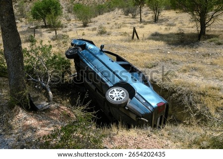 Dramatic image of a blue car overturned in a dry roadside ditch after an accident on a rural road. The vehicle appears damaged and abandoned in a remote, grassy area surrounded by trees and bushes.