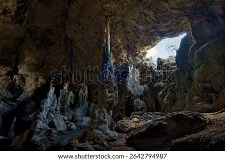 Dramatic interior of a natural cave in Spain, featuring striking stalagmites and stalactites illuminated by natural light pouring in through a large opening in the cave ceiling. Underground landscape.
