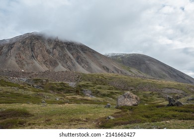 Dramatic landscape with big stones on hills against high mountain with sharp rocks in rainy low clouds. Bleak scenery with large rocky mountain range in low gray cloudy sky in gloomy rainy weather.