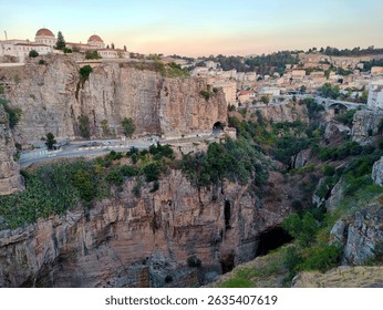 Dramatic landscape of Constantine, Algeria, during the golden hour light. It showcases the deep gorges carved by the Rhumel River, iconic limestone cliffs, the Bab Al-Qantara Bridge in the distance.