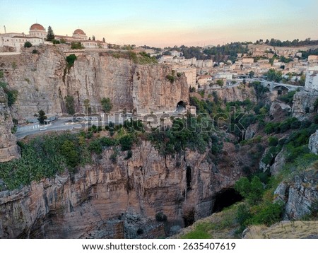 Dramatic landscape of Constantine, Algeria, during the golden hour light. It showcases the deep gorges carved by the Rhumel River, iconic limestone cliffs, the Bab Al-Qantara Bridge in the distance.