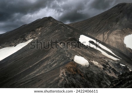 Dramatic landscape of dark rugged volcano with moody sky and hiker hiking on Blahnukur trail among the Icelandic Highlands in summer at Landmannalaugar, Iceland