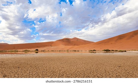 A dramatic landscape featuring a cracked, parched dry lake bed in the foreground, leading toward massive orange sand dunes under a beautiful sky