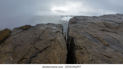 A dramatic landscape featuring a large rock formation with a deep crevice, surrounded by mist and fog, creating a mysterious and moody atmosphere. - Powered by Shutterstock