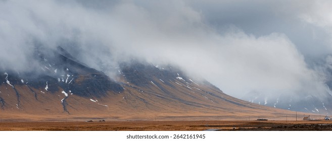 Dramatic landscape featuring low clouds obscuring majestic mountains in iceland, with small houses nestled in the foreground. Icelandic landscape