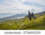 Dramatic landscape featuring the Old Man of Storr rock formation on the Isle of Skye, Scotland. Lush green hills lead to the iconic pinnacle, with a scenic coastline and distant mountains in the background. A winding path invites exploration.