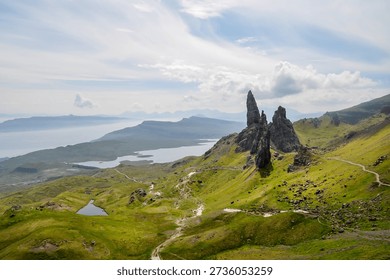 Dramatic landscape featuring the Old Man of Storr rock formation on the Isle of Skye, Scotland. Lush green hills lead to the iconic pinnacle, with a scenic coastline and distant mountains in the background. A winding path invites exploration.