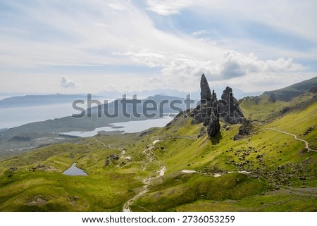 Dramatic landscape featuring the Old Man of Storr rock formation on the Isle of Skye, Scotland. Lush green hills lead to the iconic pinnacle, with a scenic coastline and distant mountains in the background. A winding path invites exploration.
