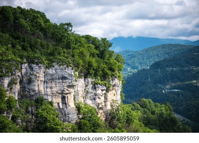 a dramatic landscape featuring a rugged cliff adorned with greenery, overlooking a valley with scattered houses amidst rolling, forest-covered hills under a moody sky.