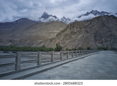 A dramatic landscape featuring rugged peaks partially covered by clouds, viewed from Machulo RCC Bridge in the Masherbrum region of Ghanche District, Gilgit Baltistan, Pakistan. A scenic mountain road and bridge lead toward the Karakoram landscape under a moody evening sky.