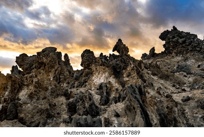 A dramatic landscape featuring rugged volcanic rock formations under a colorful sky at sunset. The scene captures the textures and colors of the rocks contrasting with the soft hues of the clouds.