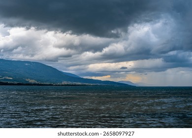 A dramatic landscape featuring a stormy sky over a Lac Neuchatel lake. Dark clouds loom above, casting shadows on the water, while distant mountains create a serene backdrop.