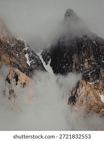 A dramatic landscape featuring towering mountain peaks shrouded in mist and partially covered with snow. The rugged terrain and steep cliffs highlight the raw beauty of nature.