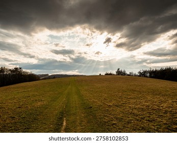 Dramatic landscape with heavenly sun rays breaking through clouds over rolling meadow and distant hills, creating divine light beams and atmospheric sky.
