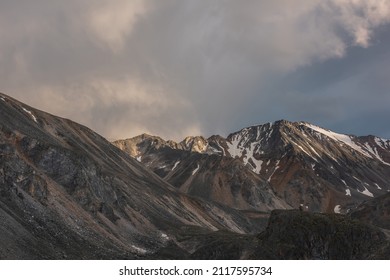 Dramatic landscape with high mountain range with sunlit golden sharp rocky top under clouds of sunset color in gloomy sky. Dark atmospheric scenery with large mountains with snow at changeable weather - Shutterstock ID 2117595734