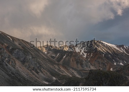 Dramatic landscape with high mountain range with sunlit golden sharp rocky top under clouds of sunset color in gloomy sky. Dark atmospheric scenery with large mountains with snow at changeable weather
