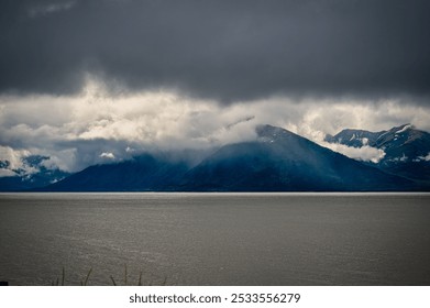 A dramatic landscape of a mountain range shrouded in clouds over a calm body of water. - Powered by Shutterstock