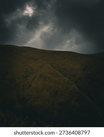 A dramatic landscape photograph featuring a dark, moody hillside under a cloudy, stormy sky. The rugged terrain is covered in muted green and brown vegetation, while the heavy clouds partially reveal a soft glow of light breaking through. The contrast between the shadowed slopes and the turbulent sky creates an atmospheric, cinematic scene. The image evokes themes of wilderness, mystery, solitude, and raw natural beauty.