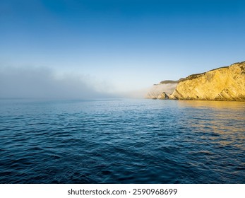 A dramatic landscape of a rocky cliff side on a cloudy day, with the misty ocean stretching out in the foreground