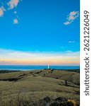 Dramatic landscape of rolling grasslands sloping towards the vast blue ocean under a soft twilight sky, featuring a striking lighthouse as a focal point of coastal beauty. April 25, 2025. Savu Island.