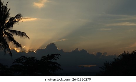 Dramatic landscape with sun rays piercing through thick dark clouds over a tropical forest and distant mountains. Ideal for themes of hope, peace, nature, and spirituality.