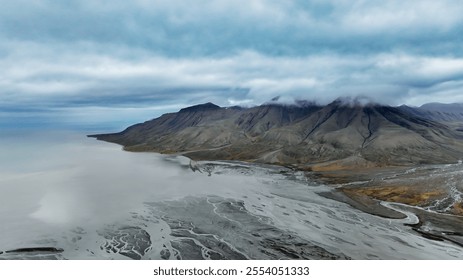 The dramatic landscape of Svalbard features a rugged coastline meeting the sea, with mountains shrouded in clouds. The scene captures the raw beauty of this Arctic wilderness during a cloudy day.