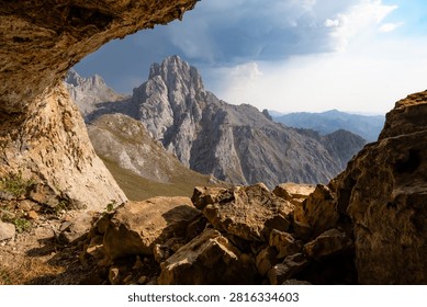 A dramatic landscape viewed from inside a cave toward a rugged mountain peak in Picos de Europa National Park, Spain. A towering thunderstorm cloud illuminated by the warm light of the setting sun creates a striking contrast between light and shadow in this wild mountain scenery.