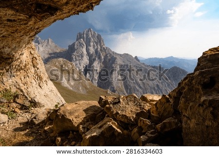 A dramatic landscape viewed from inside a cave toward a rugged mountain peak in Picos de Europa National Park, Spain. A towering thunderstorm cloud illuminated by the warm light of the setting sun creates a striking contrast between light and shadow in this wild mountain scenery.